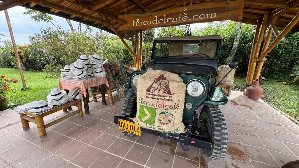 Un coche antiguo estacionado bajo un refugio de madera con una mesa llena de sombreros y un cartel que dice "fincadelcafe.com". 3