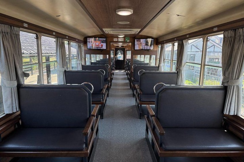 Interior de un tren con hileras de asientos, ventanas y una pantalla de televisión. 3