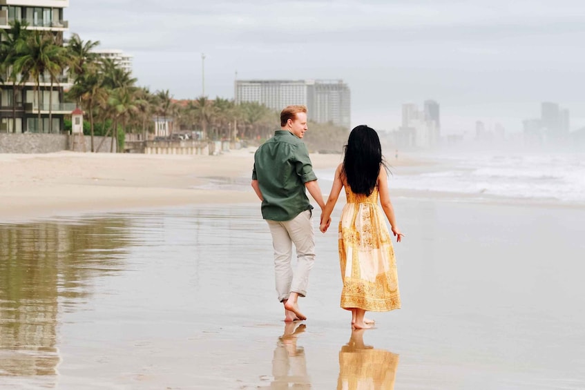 A couple walking hand in hand on a beach with palm trees and buildings in the background. 2