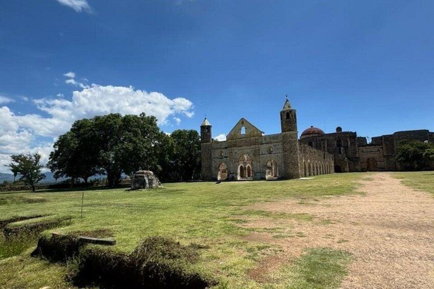 A historic stone building with a bell tower, surrounded by a grassy field and trees. 2
