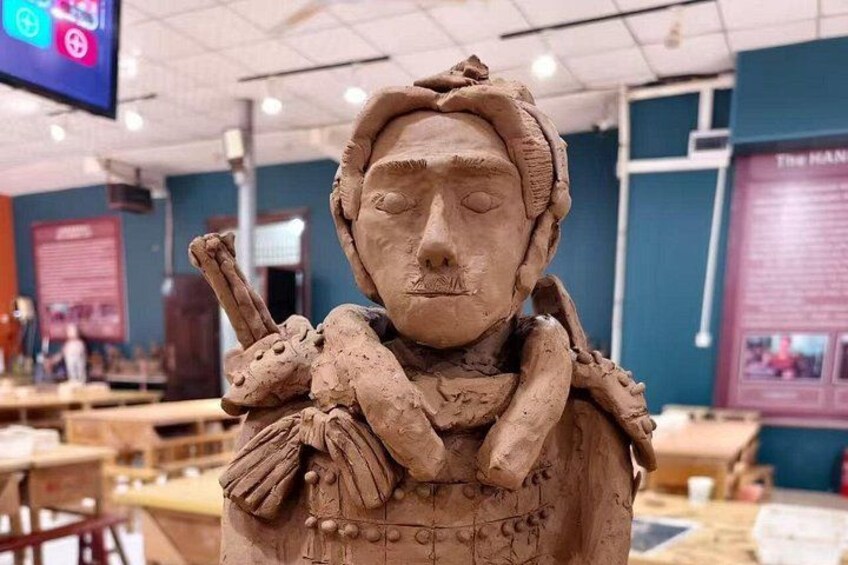 En skulptur i lera som föreställer en figur med ett pannband, som håller i en stav och bär en klädnad med ett mönster. 4