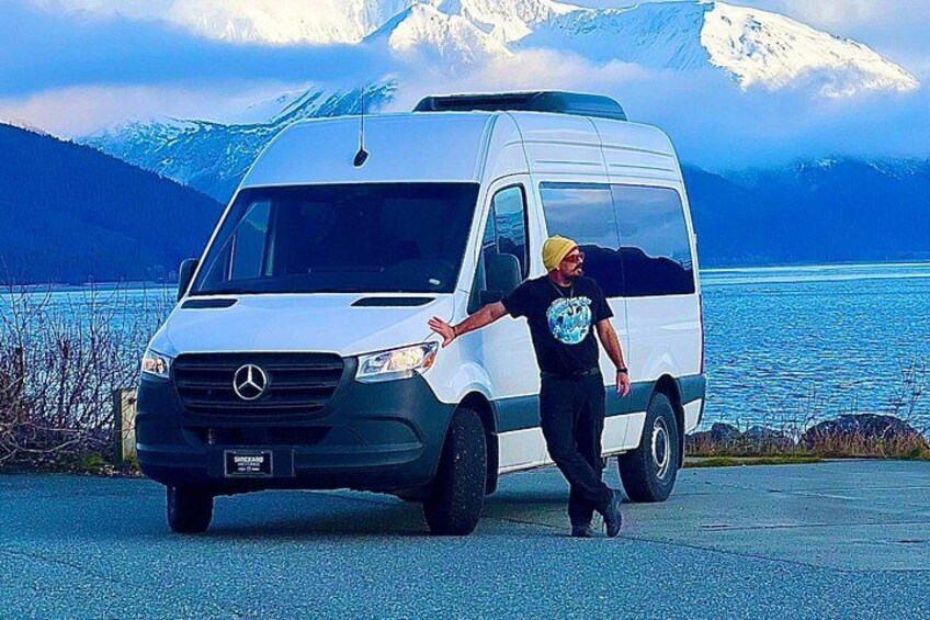 Ein weißer Van steht an einer Straße, neben ihm eine Person, im Hintergrund ein See und schneebedeckte Berge. 2