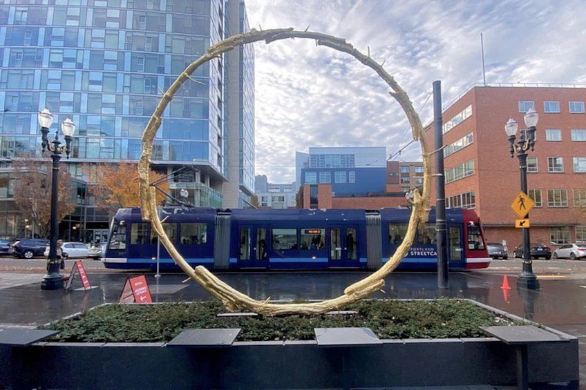 Una escultura circular en un entorno urbano, con edificios y un tranvía al fondo. 1