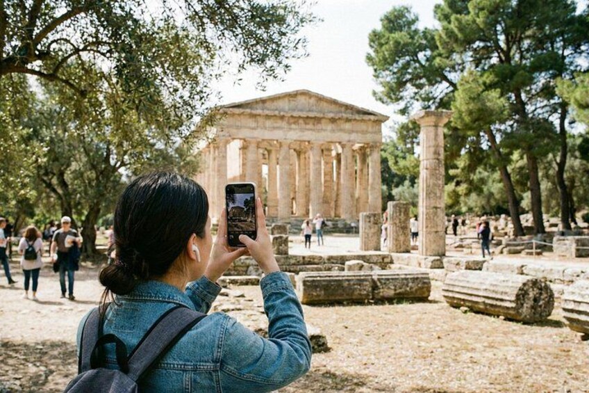 A person taking a photo of ancient ruins with columns and a temple-like structure in the background. 4