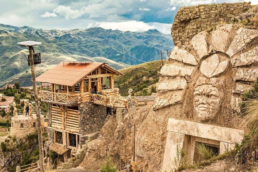 Una estructura de madera con techo rojo, un muro de piedra con rostro tallado y un paisaje montañoso al fondo. 1