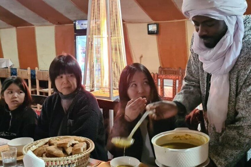 A person serving food to two women at a table with a basket of bread and a pot of soup. 4