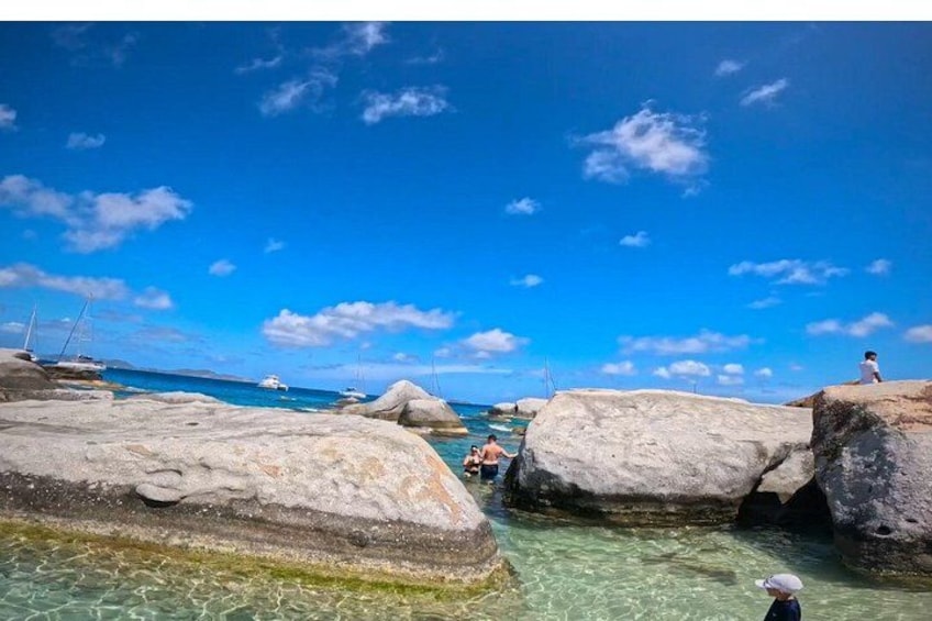 A rocky shoreline with clear water, boats in the distance, and a person walking. 5
