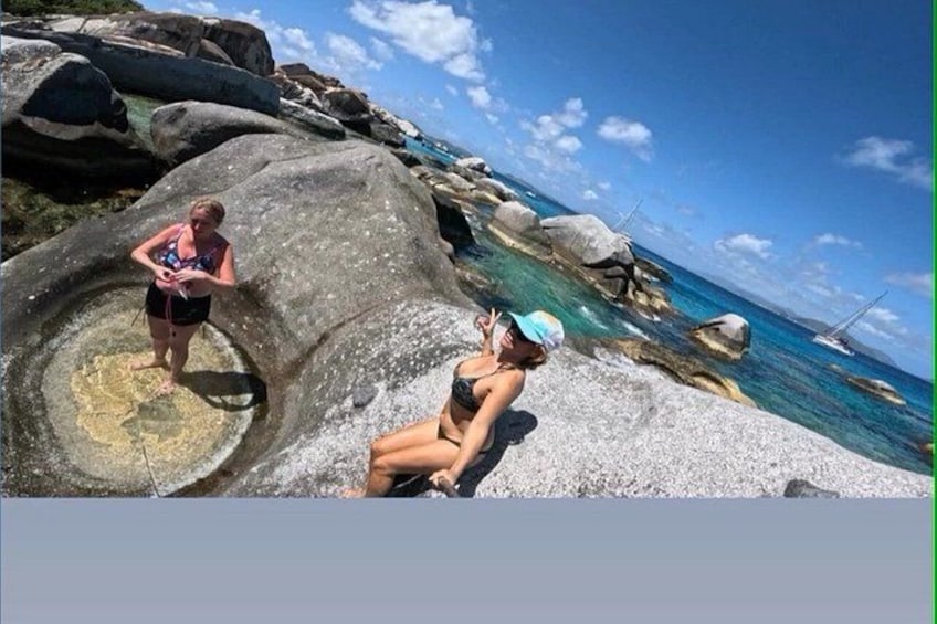 Two people near a rocky shoreline with clear blue water and a sailboat in the distance. 2