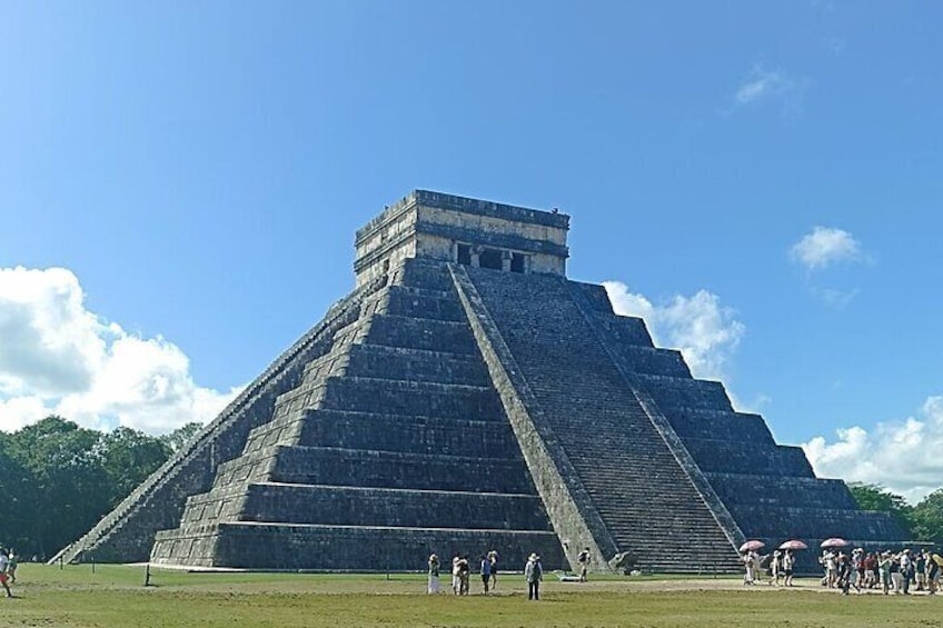 Une grande structure pyramidale à étages, entourée d’une zone herbeuse et surplombée d’un ciel bleu clair. 1
