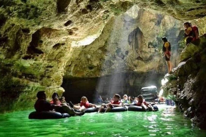 People floating in a cave on green water, with rocky walls and a person standing on a rock. 1