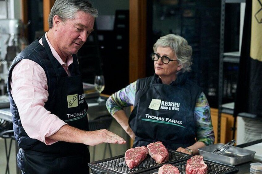 Two people wearing aprons with 'REURIEU FOOD & WINE' and 'THOMAS FARMS' logos, preparing meat on a grill. 3