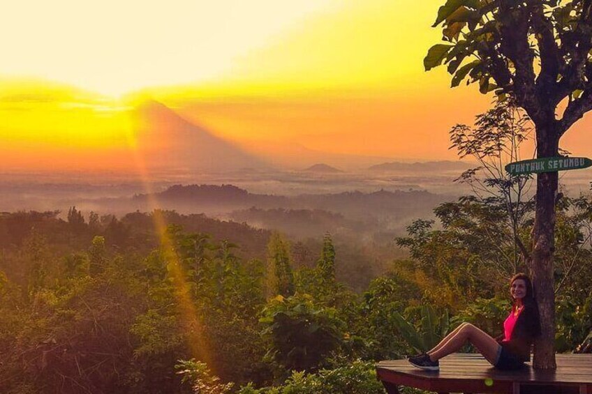 A person sitting on a wooden platform with a sign reading "PUNTHUK SETANBU" in a forested area during sunset. 1