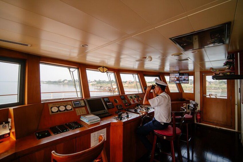 Ponte di comando di una nave con una persona al timone, diverse console di controllo e finestre vista sull'acqua. 3