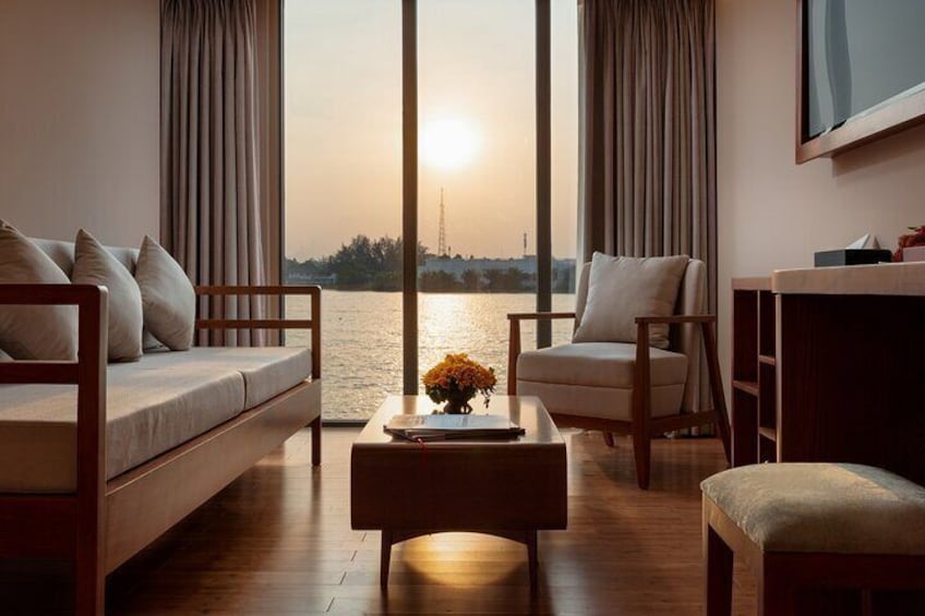 A living room with a sofa, armchair, and ottoman, a coffee table with flowers, and a large window overlooking a body of water at sunset. 3