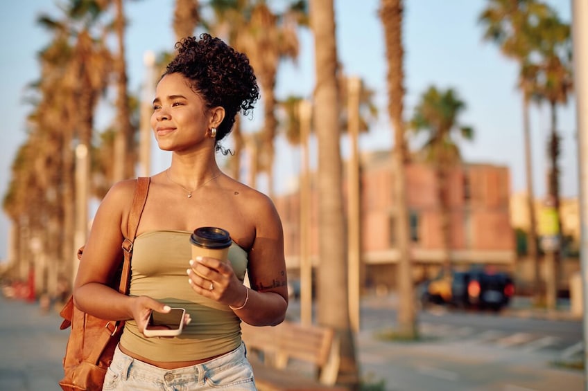 Una persona sosteniendo una taza de café y un teléfono inteligente, de pie en una acera con palmeras y edificios al fondo. 5