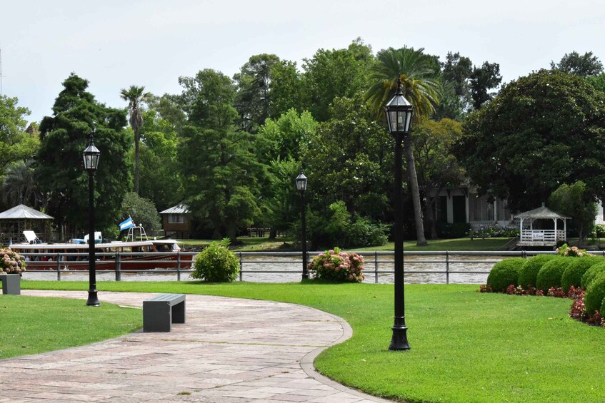 A park with a paved walkway, benches, lamp posts, and a boat on the water. 5