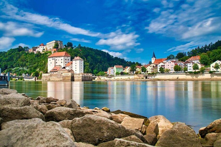 A calm river with a castle on a hill, surrounded by buildings and greenery. 1