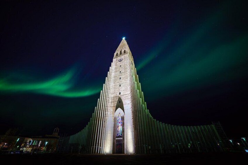 Un edificio alto e iluminado con techo puntiagudo y una gran entrada arqueada, frente a un cielo nocturno con aurora boreal verde. 2