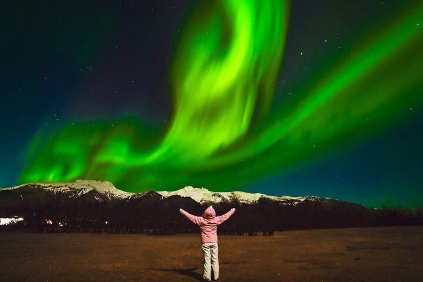 Una persona de brazos alzada, mirando hacia una cordillera bajo un cielo nocturno con auroras boreales verdes. 3