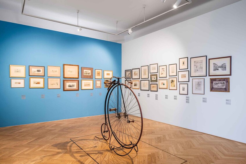 Ett konstgalleri med inramade konstverk på blå och vita väggar, och en gammal cykel placerad i mitten. 4
