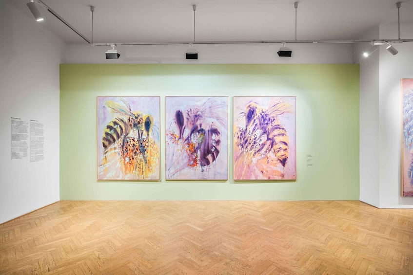 An art gallery with three abstract paintings on a light green wall and wooden parquet flooring. 2
