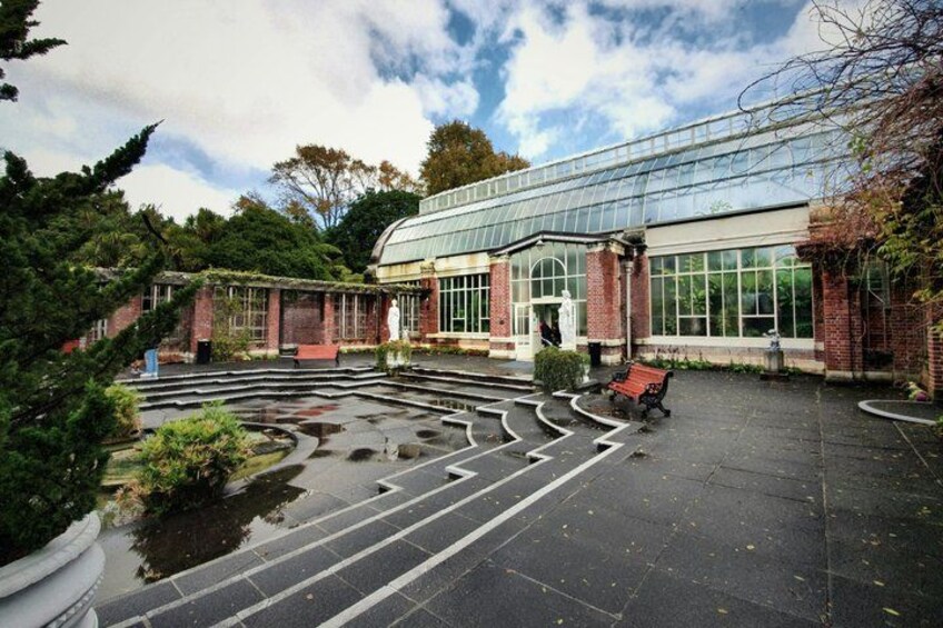 A greenhouse with a glass roof, surrounded by a brick building, a wet pavement, and a bench. 5