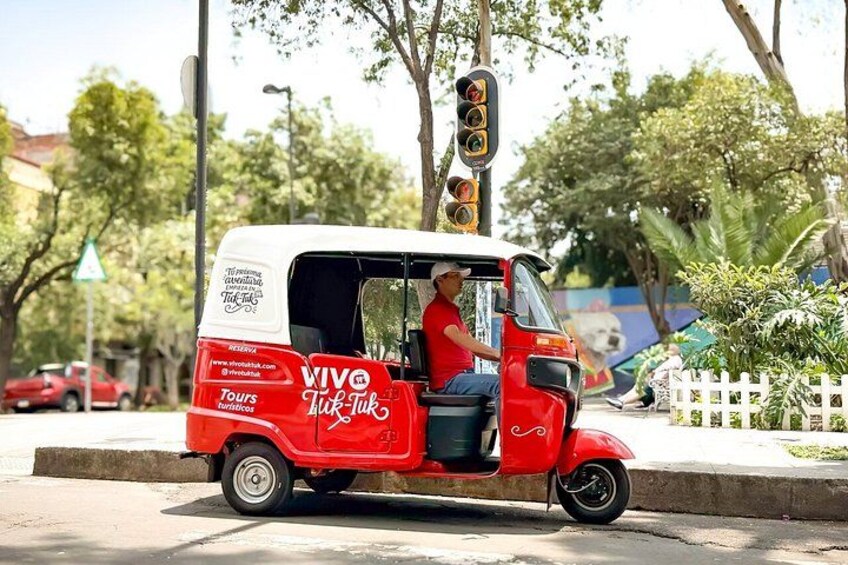 En röd, automatisk trehjulig taxi med "VIVO Tuk Tuk"-logotyp, ett trafikljus och träd i bakgrunden. 3