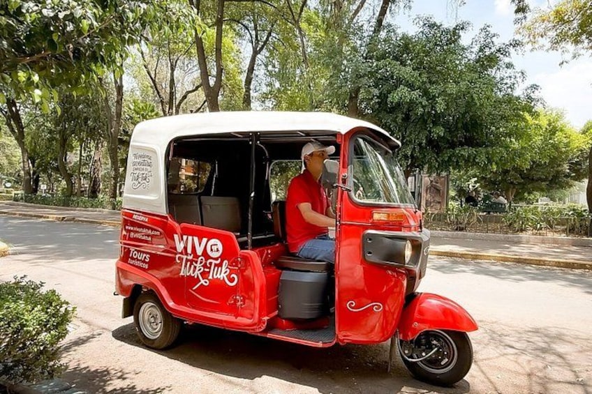 En röd, automatisk rickshaw står parkerad på en väg med träd i bakgrunden. 1
