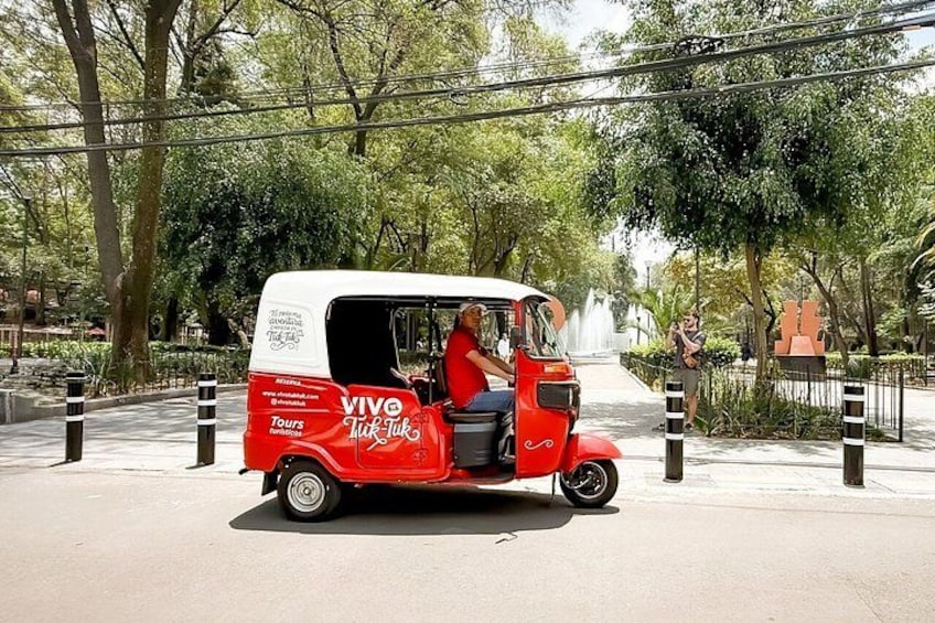 En röd och vit tuk-tuk med orden ”VIVE” och ”Tours” målade på sidan, parkerad på en gata med träd och en fontän i bakgrunden. 2
