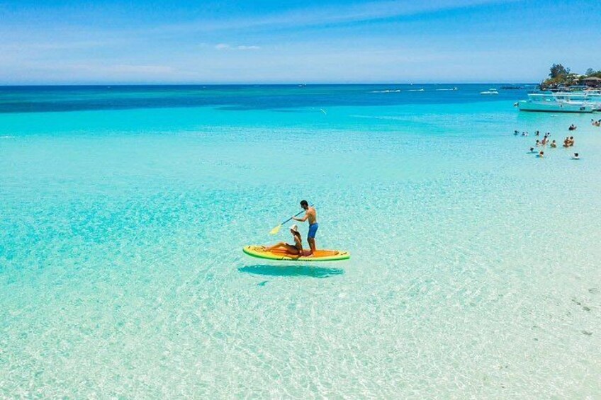A person paddleboarding on a clear turquoise sea with a sandy beach and distant boats. 5