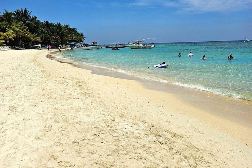 A sandy beach with clear turquoise water, palm trees, and boats in the distance. 2