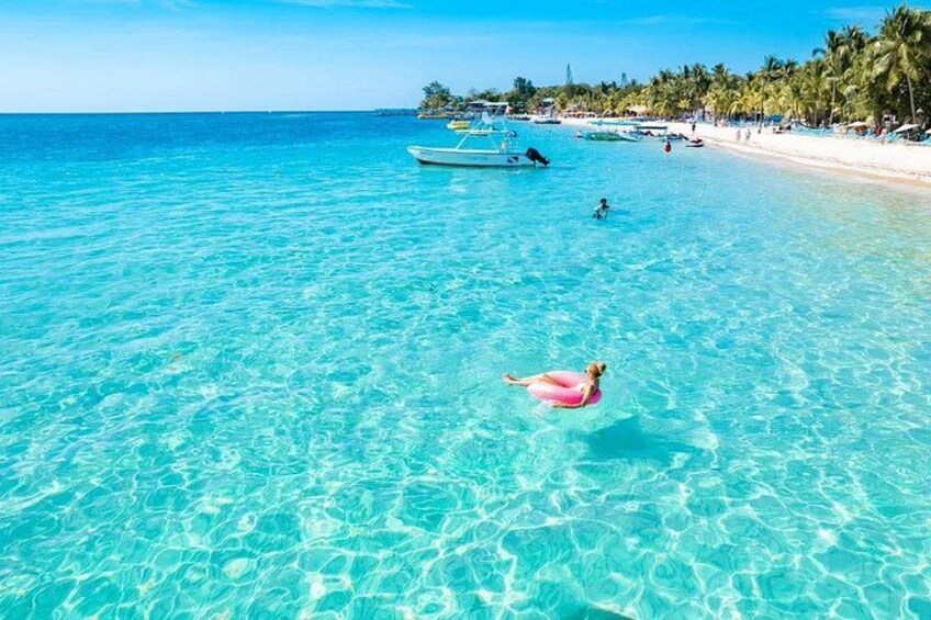 A clear turquoise sea, a person floating on a pink float, and a beach with palm trees and boats in the background. 4
