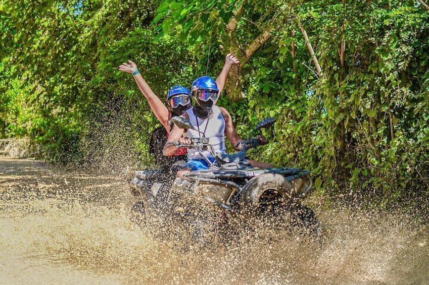 Two people on an ATV driving through a muddy area with trees in the background. 1