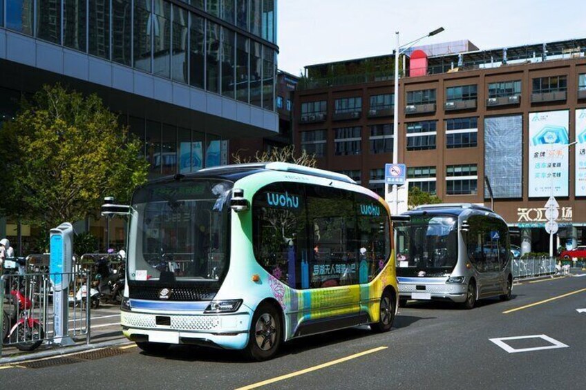 Een moderne stadstraat met twee bussen, waarvan er één het logo 'Wohu' heeft, en een gebouw met grote ramen op de achtergrond. 2