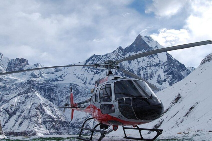 En helikopter med røde og hvide farver, parkeret på en snedækket bjergskråning med sneklædte tinder i baggrunden. 2