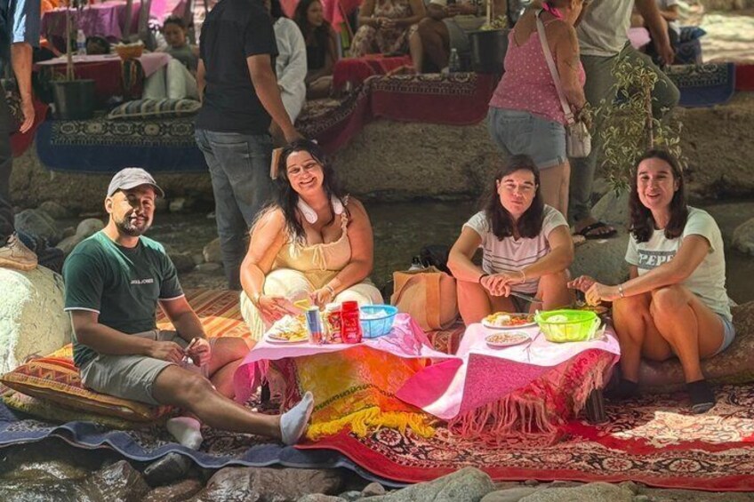 People sitting on a mat by a river, with a table in front of them. 4
