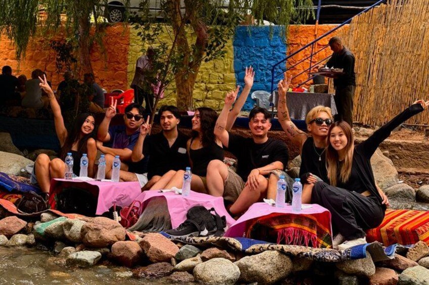 Un grupo de personas sentadas sobre rocas junto a un arroyo, con botellas de agua y un mantel rosa. 3
