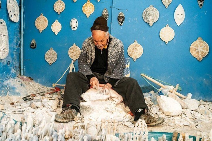 Una persona sentada en el suelo, rodeada de fragmentos de cerámica y herramientas, con una pared azul decorada con diversas máscaras de cerámica al fondo. 4