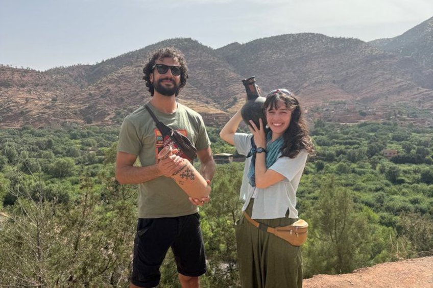 Two people holding a large clay pot, standing on a hill with a view of a green valley and hills in the background. 2