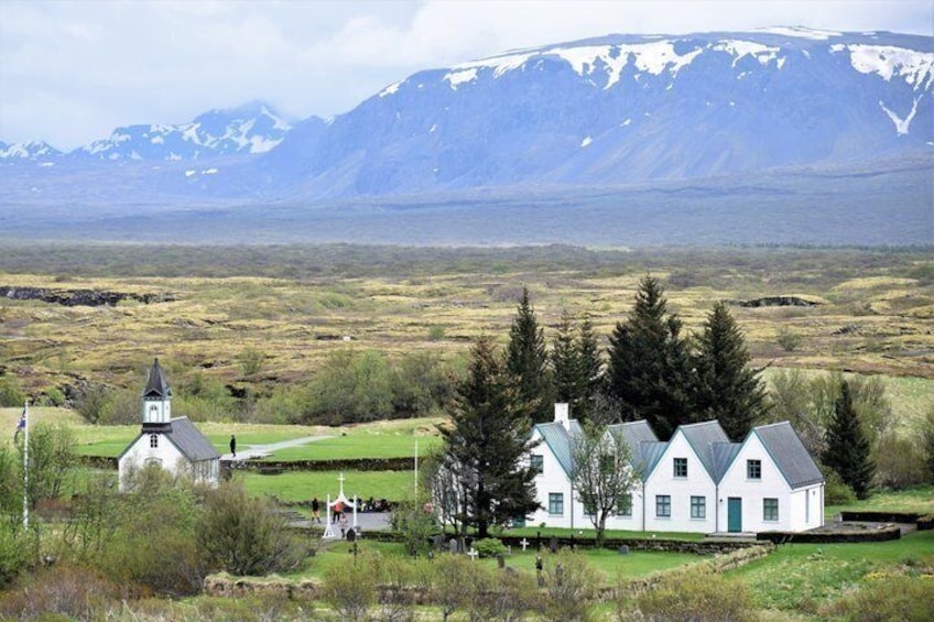 En vit kyrka med ett torn, ett vitt hus med ett grönt tak och ett bergigt landskap i bakgrunden. 3