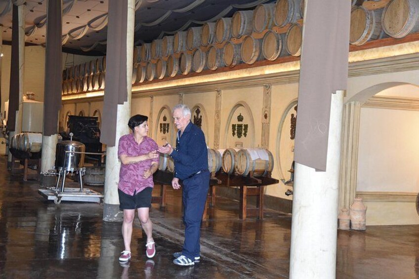 Una persona con camisa morada y shorts negros está de pie junto a un hombre con chaqueta azul y jeans. Ambos se encuentran en una habitación con barriles de madera y un contenedor metálico grande. 4
