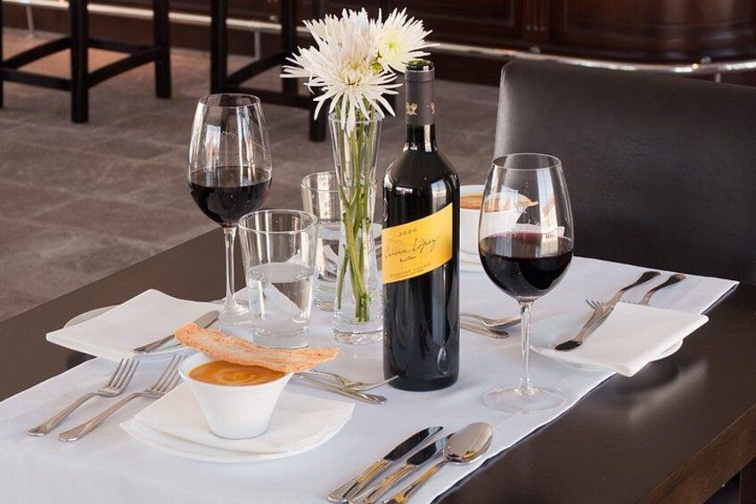 Una mesa de comedor dispuesta con copas de vino, una botella, un florero con una flor y un plato con pan. 1