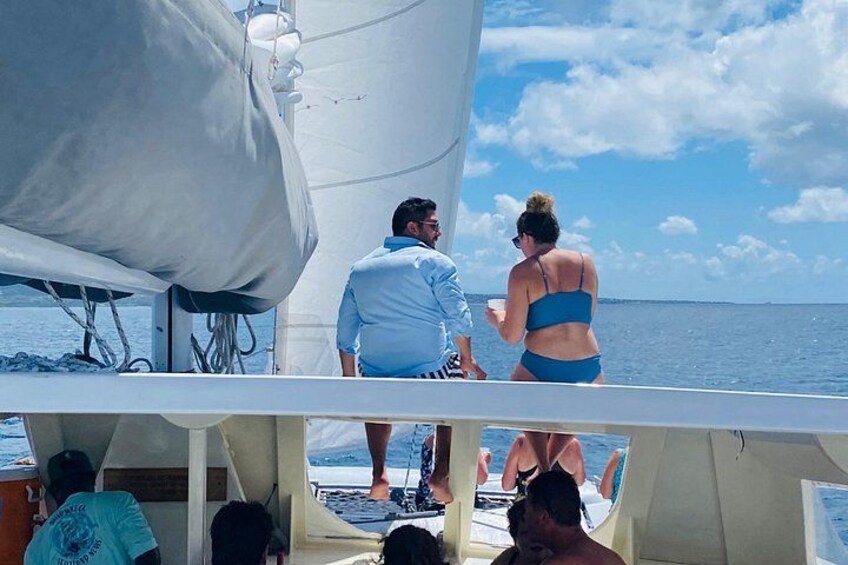 A sailboat with a person in a blue swimsuit standing at the stern, another person in a blue shirt at the bow, and a third person in a blue shirt at the stern. 2