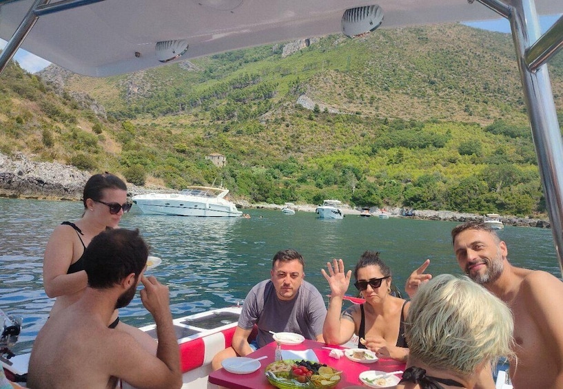 A group of people on a boat with a table of food and drinks, surrounded by water and greenery. 1