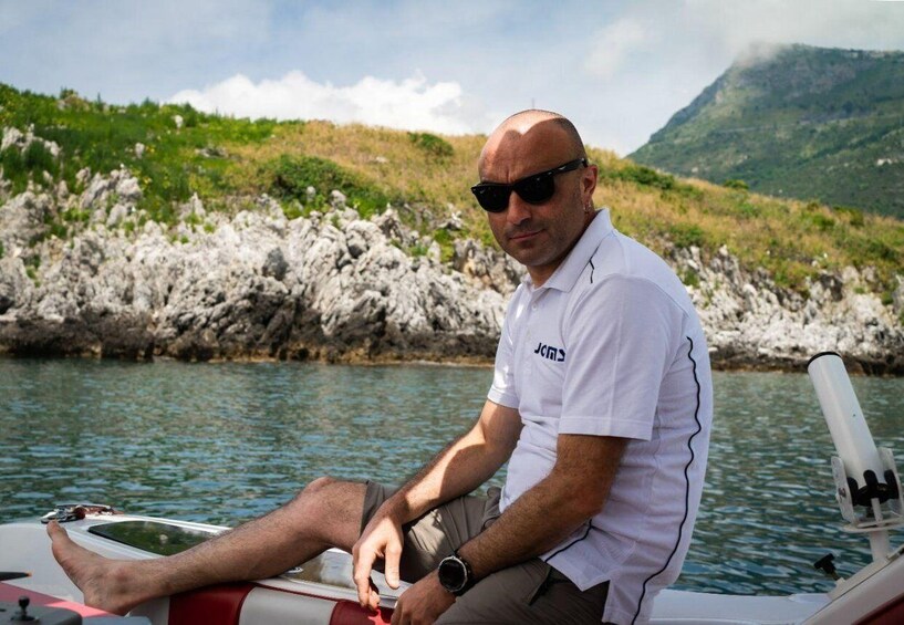 A man sitting on a boat with a rocky shoreline and a mountain in the background. 3