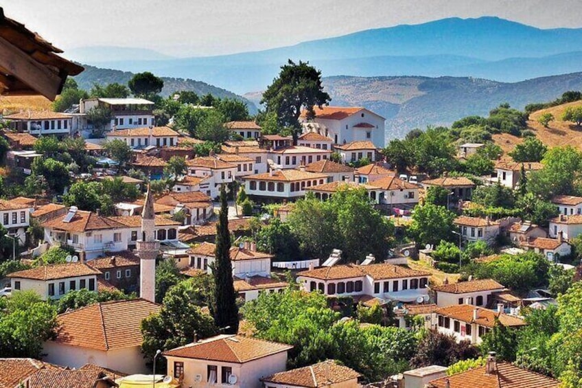 A hillside village with terracotta-roofed houses, a prominent church, and a mountain range in the background. 1