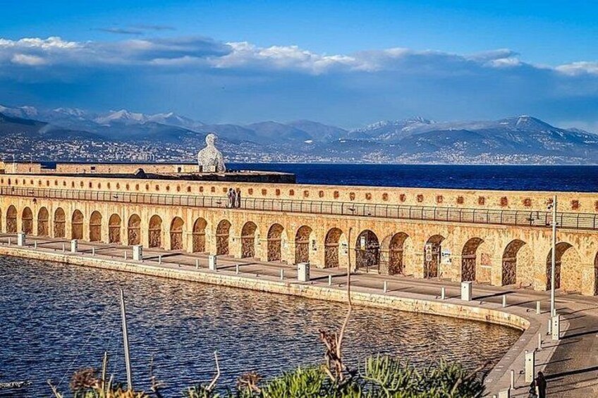A coastal walkway with arched structures, a statue, and a view of the sea and mountains in the background. 3