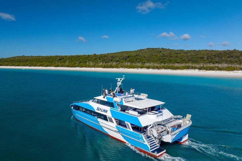 Un barco azul y blanco llamado 'SEALINK' en un mar azul despejado, con una playa y vegetación al fondo. 1