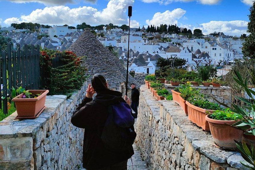 A person taking a photo of a stone wall with terracotta pots, overlooking a white village with houses and a hill in the background. 3