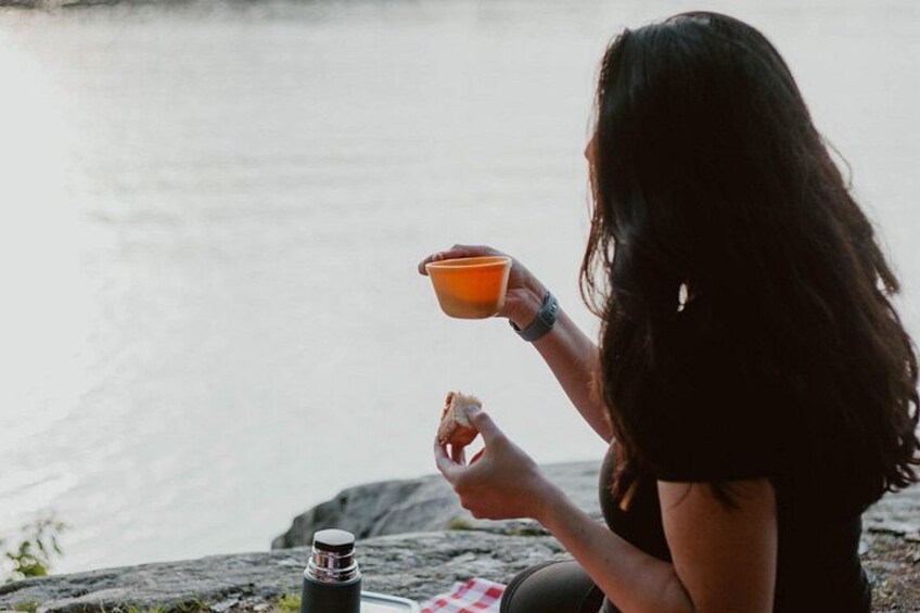 A person sitting by a body of water, holding a cup, with a bottle and a cloth nearby. 4